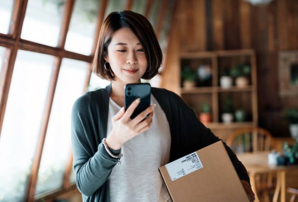 Smiling young Asian woman shopping online with smartphone on hand, receiving a delivered parcel by home delivery service. Online shopping, online banking. Enjoyable customer shopping experience